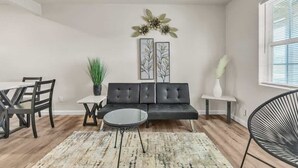 A living room with a sofa, a dining table with chairs, a coffee table, a potted plant, and artwork on the wall.