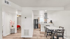 A modern kitchen with a central island, a dining area with a white table and dark chairs, and a hallway with a red bed in the background.