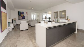 Eine moderne Küche mit einer zentralen Kochinsel, ein Essbereich mit weißem Tisch und Stühlen sowie ein Wohnzimmer mit Sofa und Couchtisch.