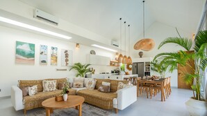Ein modernes Wohnzimmer mit einer Couch, einem Couchtisch und einem Essbereich. Es befinden sich Wandklimaanlagen, Pendelleuchten und eine große Küsterpflanze dort.