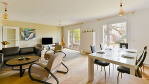 A spacious living room with a flat-screen TV, a dining area with a white table set for four, and a large window offering a view of the outdoors.