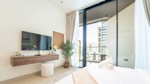 A modern hotel room with a large bed, a flat-screen TV mounted on the wall, a small table with chairs, and a balcony with a city view.