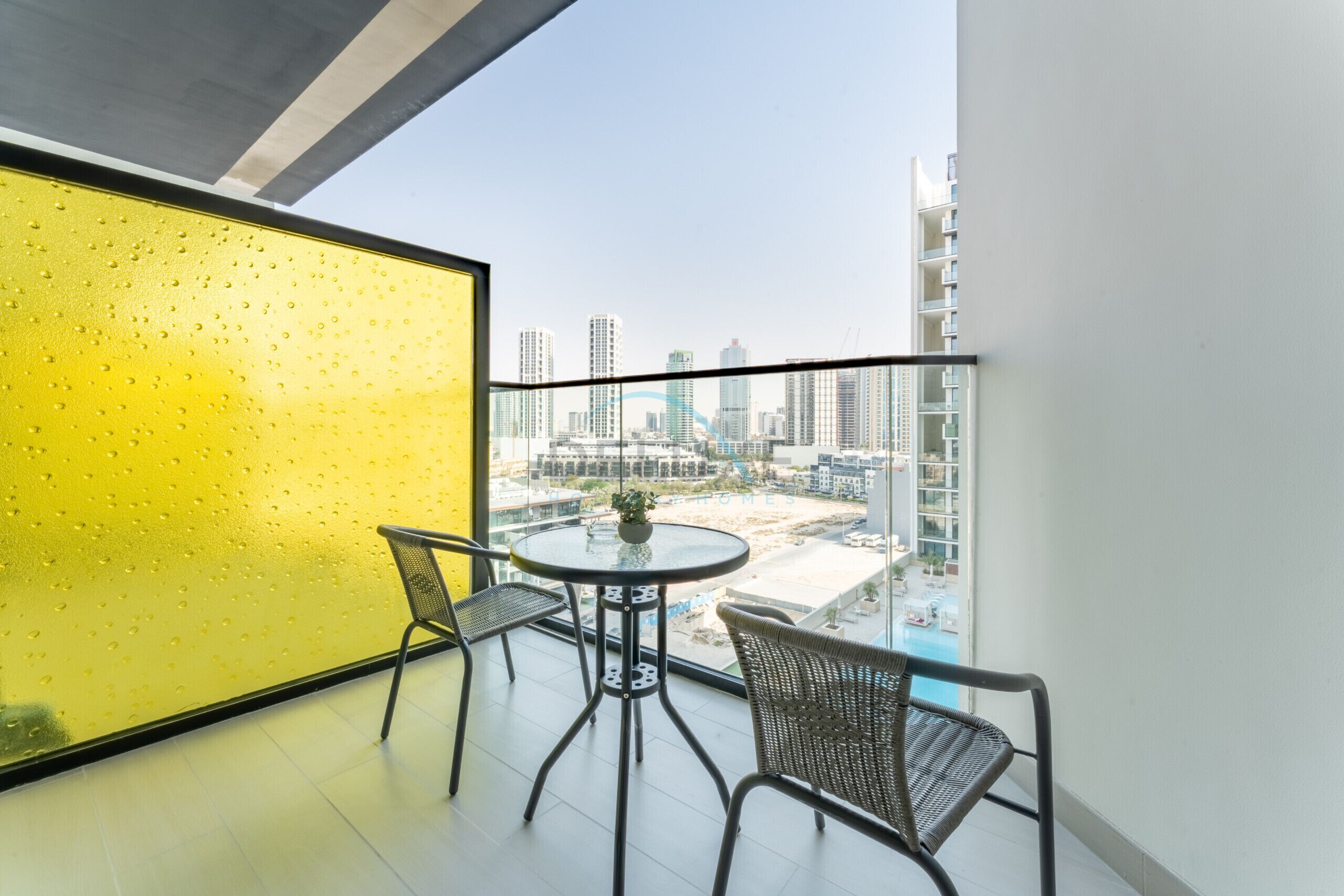 Un balcón con un muro de vidrio esmerilado amarillo, una mesa redonda con dos sillas y vistas de un paisaje urbano con edificios altos.