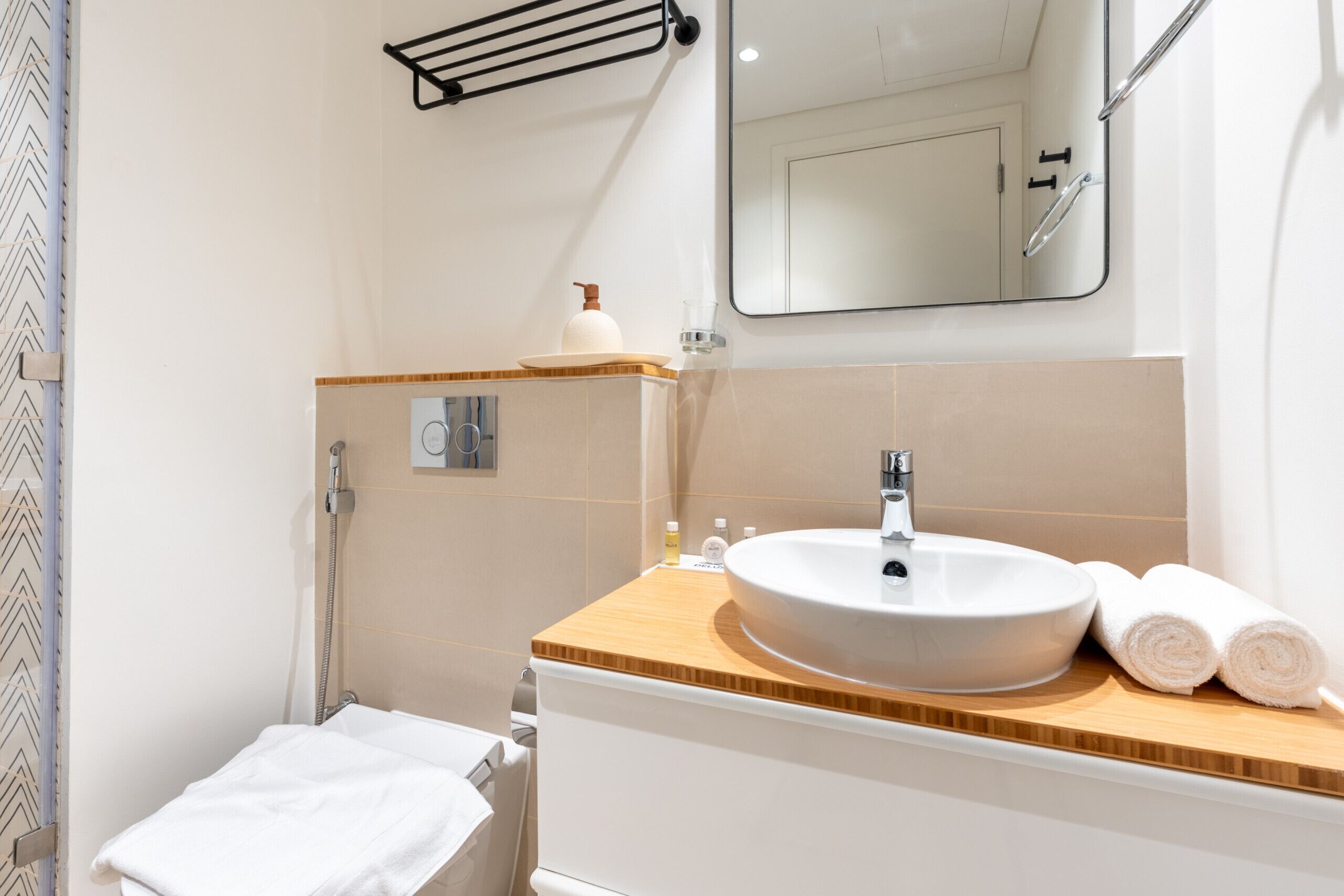 Une salle de bain moderne avec un lavabo blanc, un plan de toilette en bois et un miroir au-dessus. On aperçoit une serviette sur le sol et, au fond, l’espace réservé à la douche.