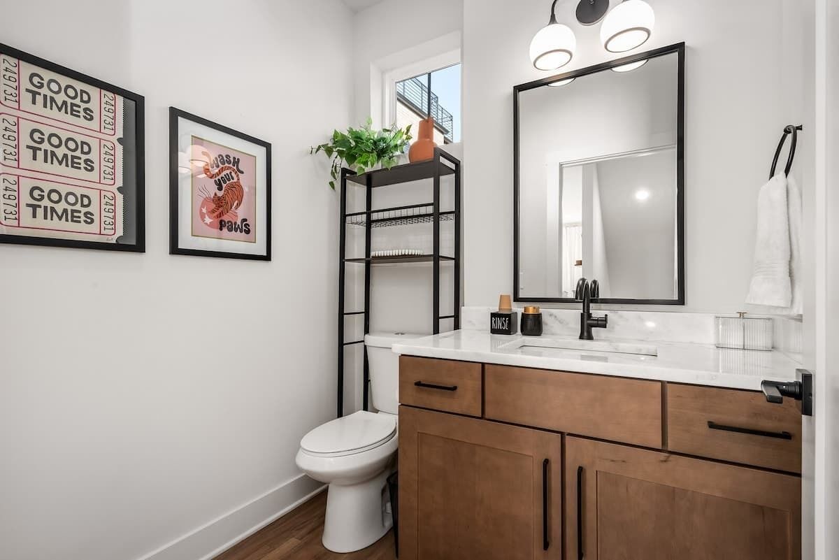 Un baño moderno con inodoro blanco, tocador de madera con espejo y lavamanos, y estante negro con una planta.