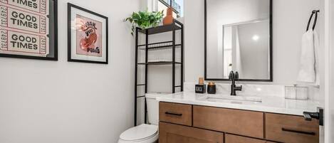 Un baño moderno con inodoro blanco, tocador de madera con espejo y lavamanos, y estante negro con una planta.