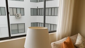Un salon moderne avec un canapé beige, une table en verre surmontée d’une lampe, et une vue sur un immeuble doté de nombreuses fenêtres.