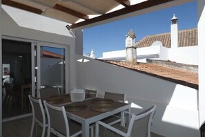 Un balcon avec une table et des chaises, une vue sur un immeuble doté d’une tour, et un ciel dégagé.