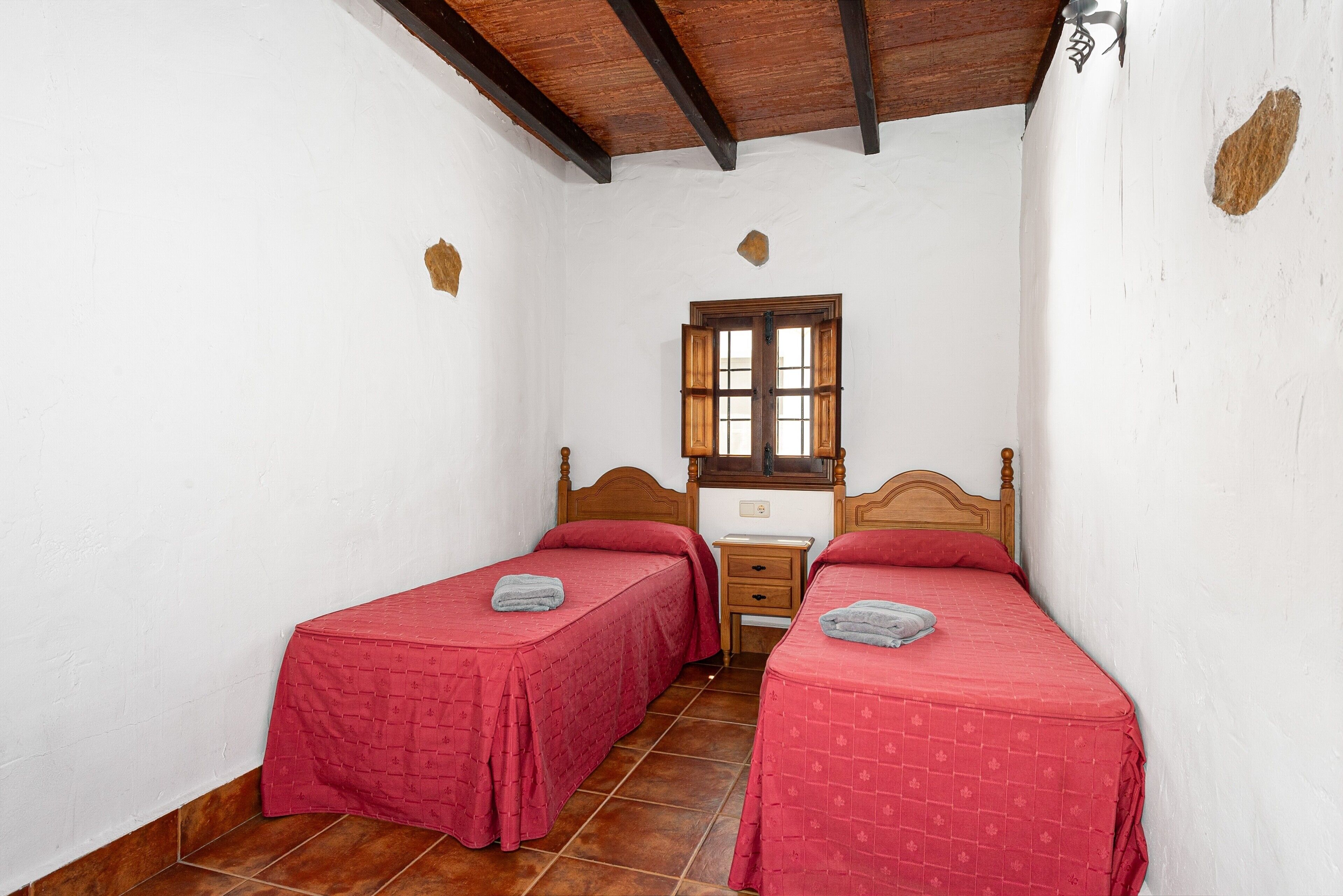 A bedroom with two single beds, a wooden headboard, a small bedside table, a window, and a white wall.