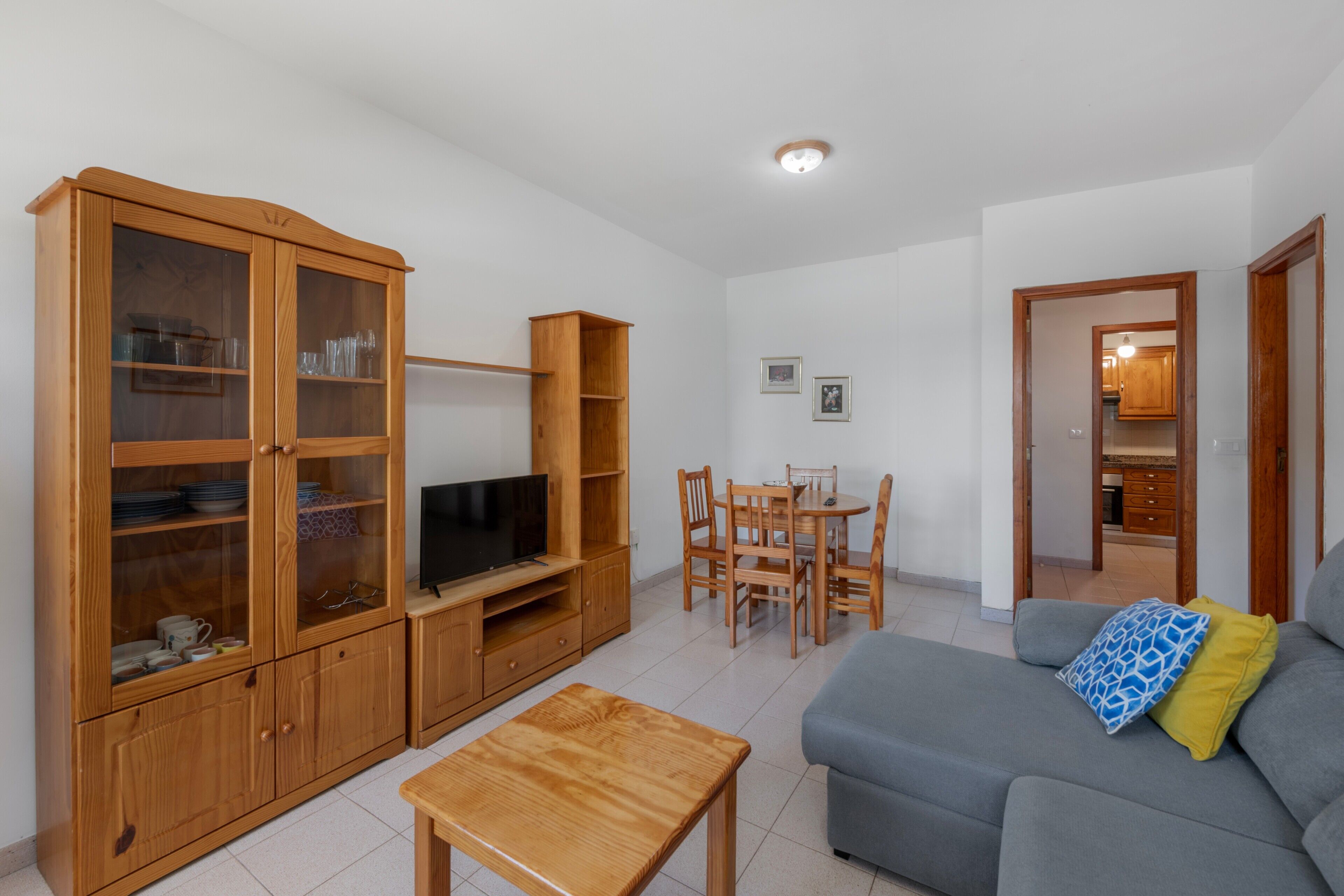 Una sala de estar con televisor, armarios de madera, una mesa de comedor con sillas y un sofá con cojines.
