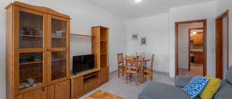 Una sala de estar con televisor, armarios de madera, una mesa de comedor con sillas y un sofá con cojines.