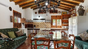 A cozy living room with a dining table, wooden chairs, a sofa, and a kitchen area with a refrigerator and shelves. - Cottage 'Majolero 2' with Shared Pool, Wi-Fi and Air Conditioning (Castillo de Locubín)