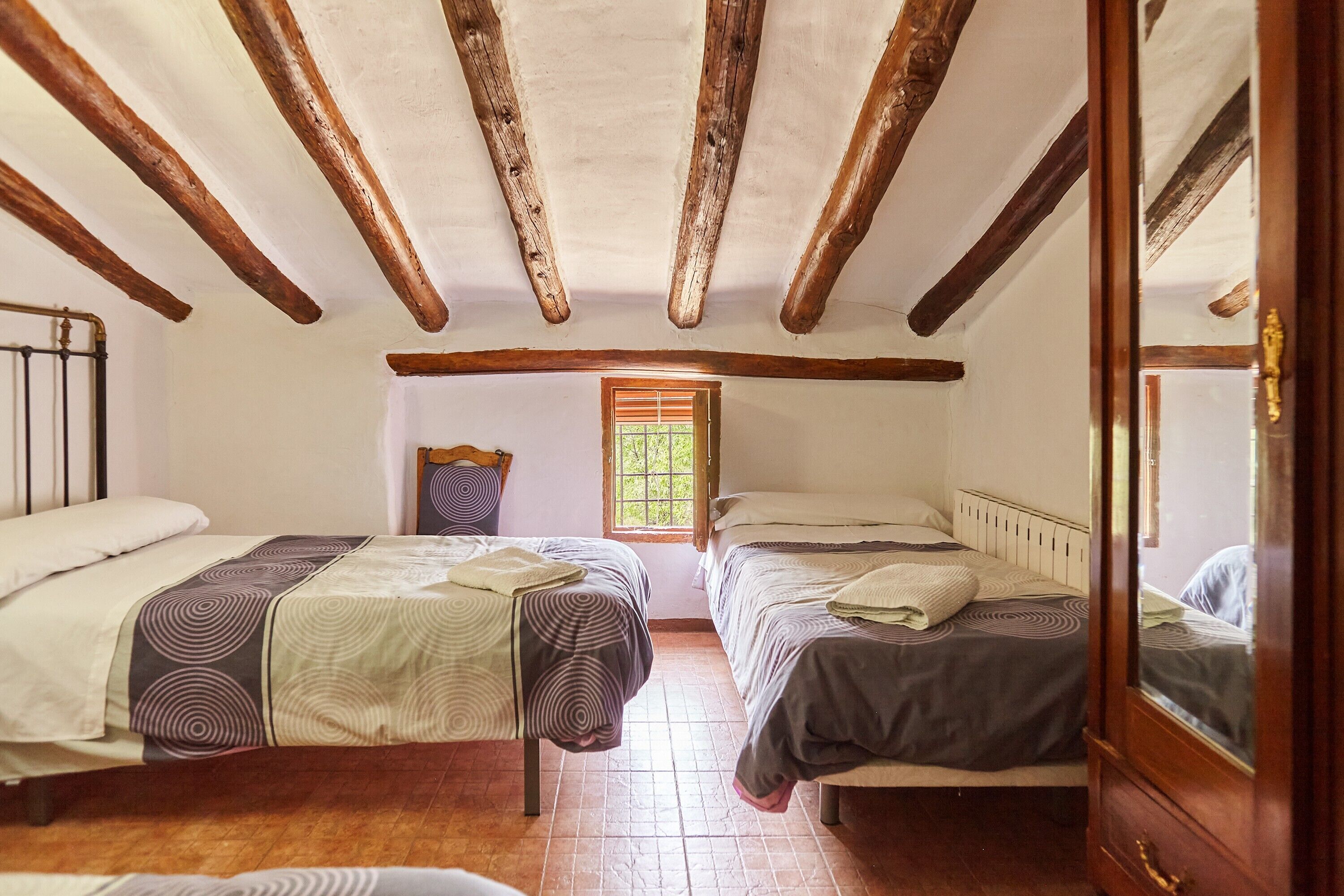Quarto com duas camas, vigas de madeira no teto, janela e porta com espelho.