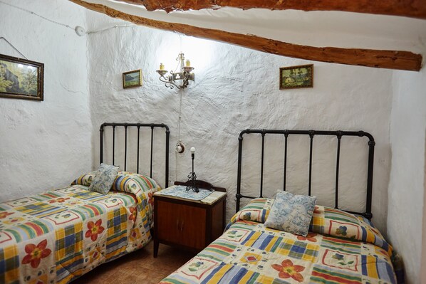 A room with two beds, a small wooden nightstand, a wall-mounted lamp, and two framed pictures.