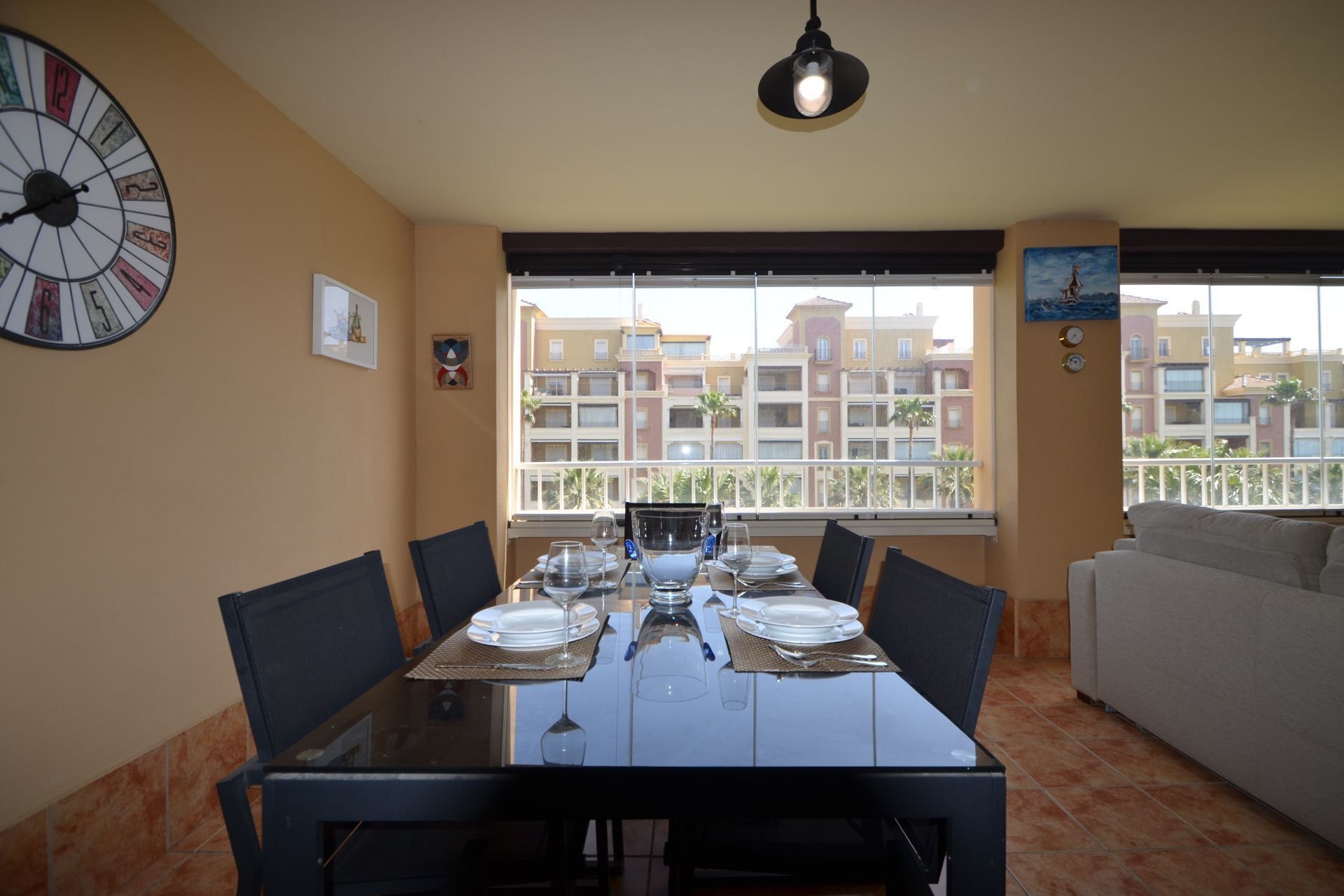A dining area with a table set for four, a large wall clock, and a view of residential buildings through large windows.