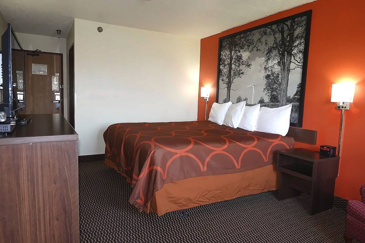 A hotel room with a bed, a desk, a chair, a nightstand, and a wall-mounted lamp.