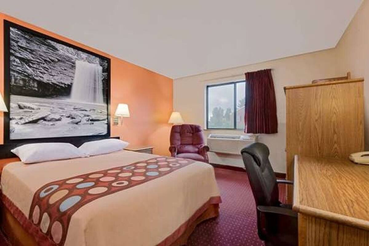 A hotel room with a bed, a desk, a chair, a window with curtains, and a wall-mounted TV displaying a waterfall.
