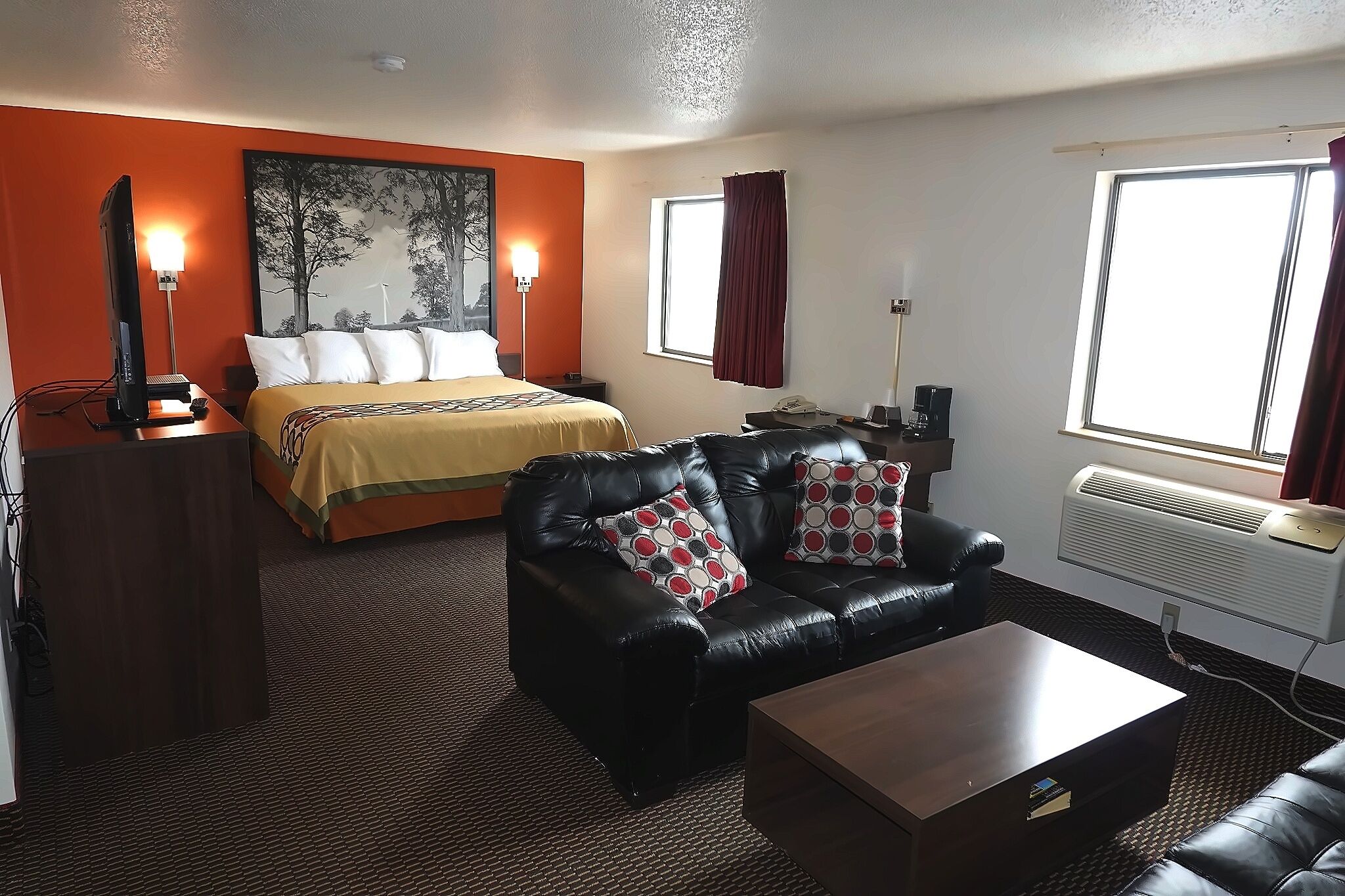 Une chambre d’hôtel avec un lit, un canapé en cuir noir, une table basse en bois, un bureau avec un ordinateur et une fenêtre avec des rideaux.
