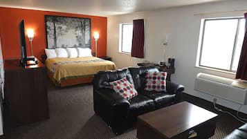 A hotel room with a bed, a black leather sofa, a wooden coffee table, a desk with a computer, and a window with curtains.
