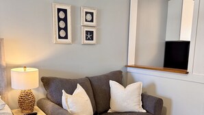 Une pièce chaleureuse avec un canapé gris, une petite table d’appoint surmontée d’une lampe, et des œuvres d’art encadrées accrochées au mur.