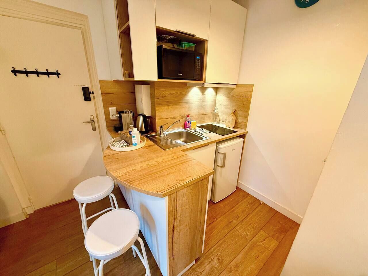 A compact kitchen with a wooden countertop, a sink, a microwave, and a small refrigerator. There are two white stools and a white door with a coat rack.