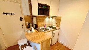 A compact kitchen with a wooden countertop, a sink, a microwave, and a small refrigerator. There are two white stools and a white door with a coat rack. - Studio 1 - Place to B "Alizé (Saint-Nazaire)