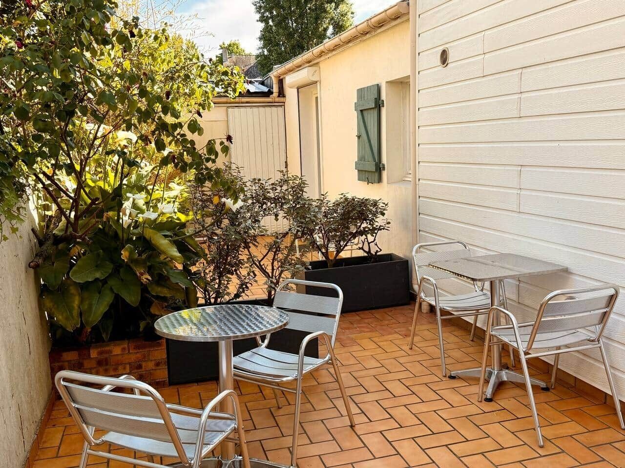 A patio with metal chairs and a round table, surrounded by potted plants and a white wall.