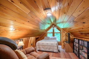 A cozy attic bedroom with a wooden sloped ceiling, a large window, a bed, a bookshelf, and a fireplace.