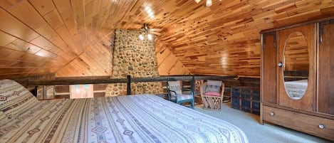 Une chambre confortable située sous les toits, avec un plafond en bois, un mur en pierre, un grand lit avec une literie à motifs, une armoire en bois et un ventilateur de plafond.