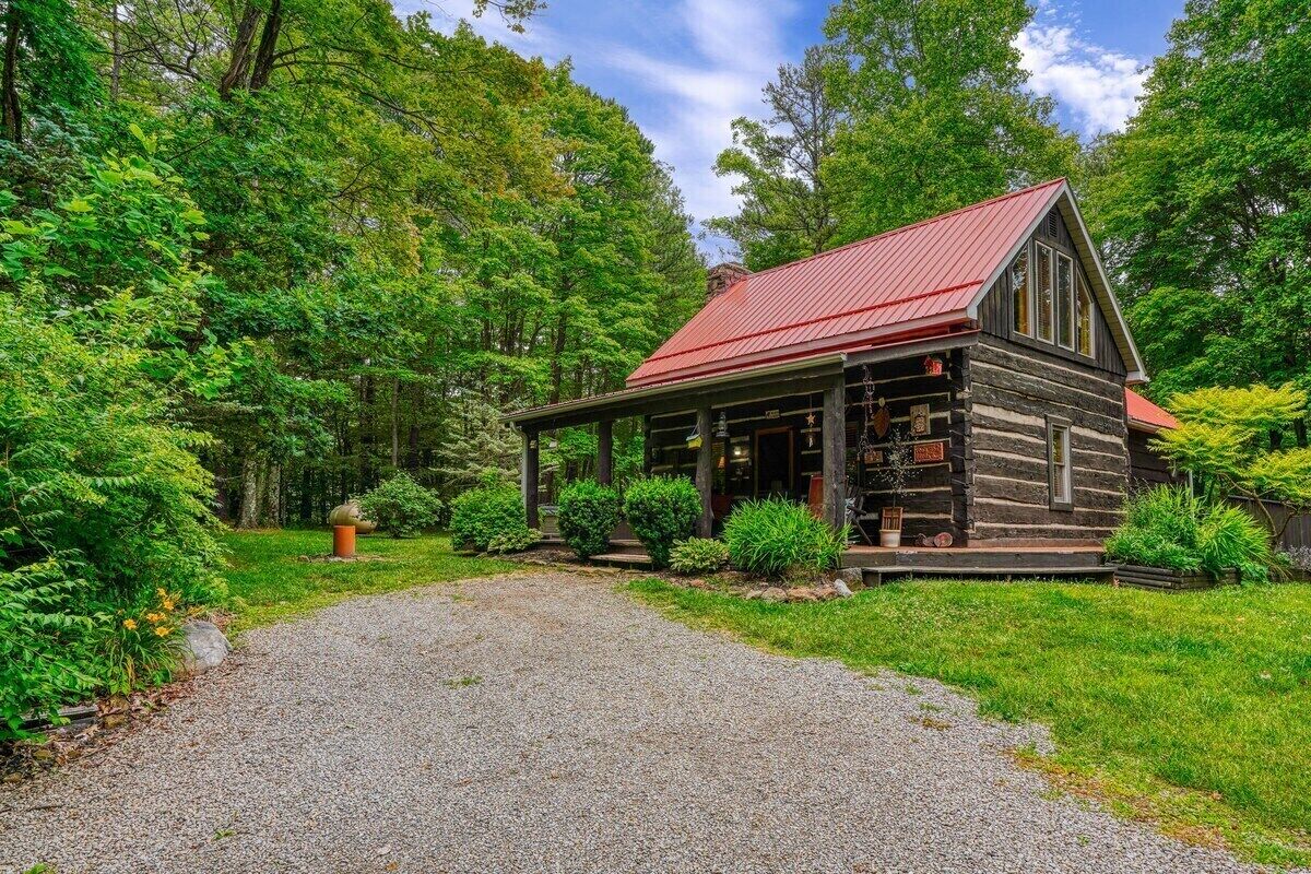 A rustic log cabin with a red roof, surrounded by lush greenery and a gravel path leading to the entrance.