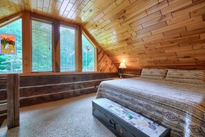 Ein gemütliches Schlafzimmer mit Holzbalkendecke, einem großen Fenster, einem Bett mit gemusterten Betttextilien und einem Koffer auf dem Boden.
