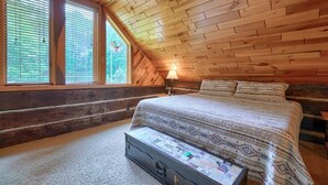 Un dormitorio acogedor en el ático, con techo inclinado de madera, una cama grande con ropa de cama estampada y una maleta en el suelo.