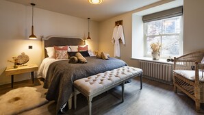 A bedroom with a large bed, bedside tables, a bench, a wicker chair, a window with a view, and a hanging robe. - Solace | Finest Stays (Totnes)