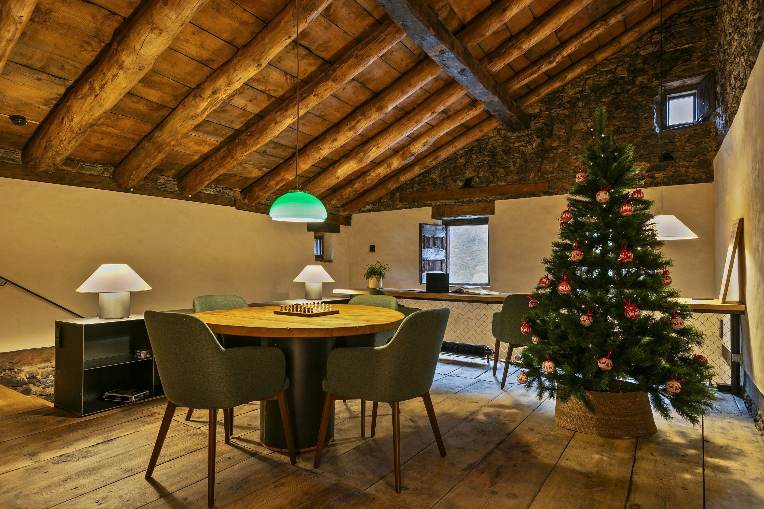 A cozy room with a wooden ceiling, a round wooden table with chairs, a Christmas tree, and two lamps.