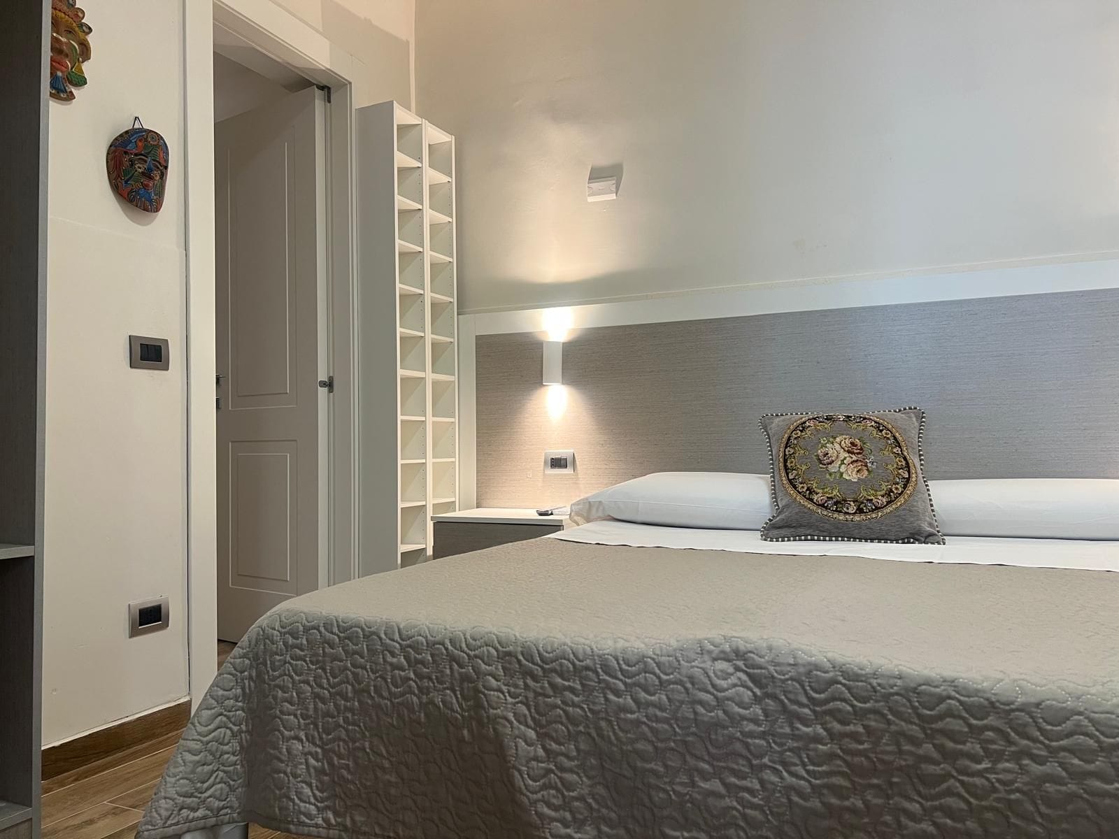 A bedroom with a bed, a nightstand, a wardrobe, and a wall-mounted shelf unit.