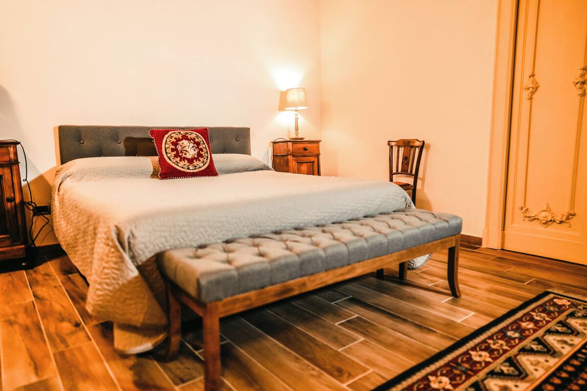 Una cama bien hecha con un almohadón rojo, una lámpara de noche y un banco de madera en una habitación con suelo de madera y alfombra estampada.