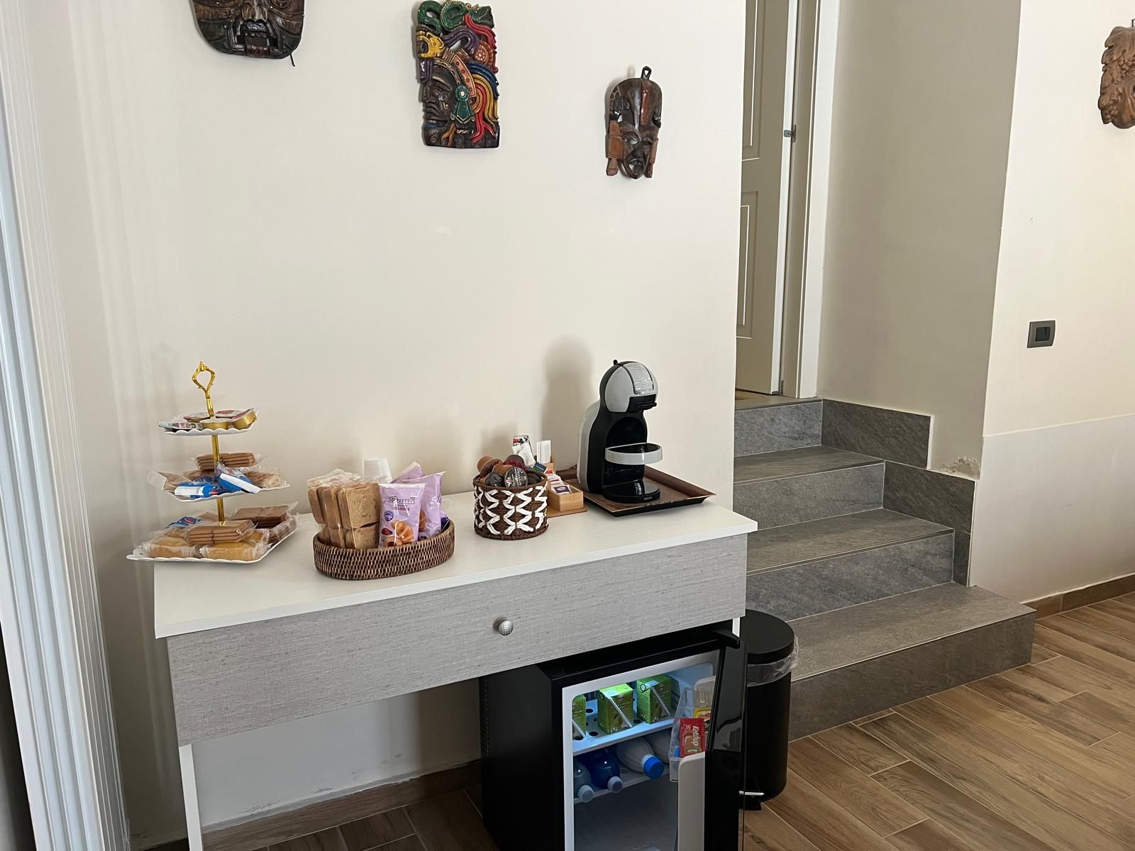 Una kitchenette con cafetera, un mini refrigerador lleno de bebidas y una bandeja de pan sobre la encimera.