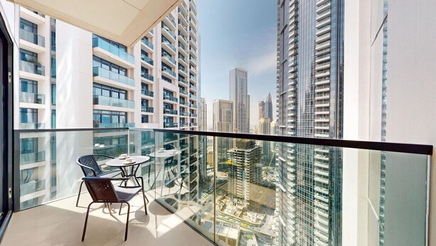 Un balcón con barandilla de vidrio, una mesa con sillas y vistas de edificios altos circundantes.