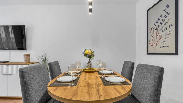 Un comedor con una mesa redonda de madera dispuesta para seis personas, sillas grises tapizadas y un jarrón con flores amarillas. Un cuadro enmarcado cuelga de la pared a la derecha.