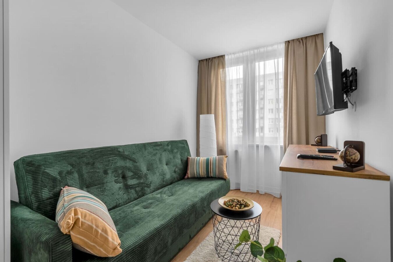 Ein modernes Wohnzimmer mit einem grünen Sofa, einem kleinen runden Tisch, einem an der Wand montierten Fernseher und einem Fenster mit durchsichtigen Vorhängen.