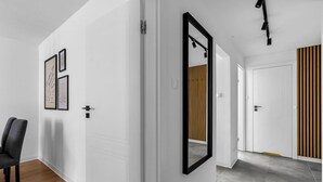 A hallway with white walls, a wooden floor, and a large mirror. There are framed pictures on the wall and a chair in the corner.