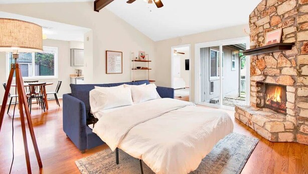 A bedroom with a stone fireplace, a ceiling fan, a bed, a blue sofa, and a dining area with a table and chairs. - 2BR 2BA Cozy Bungalow with Backyard in Montclaire - Zilker Area (Austin)