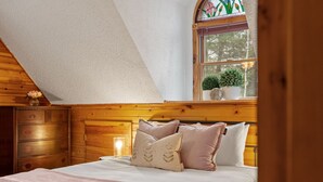 A bedroom with a sloped ceiling, wooden headboard, a bed with multiple pillows, a bedside table with a lamp, and a window with a plant. - Lucky Log Cabin- Hottub fireplace 12min to Mt Snow (Wardsboro)