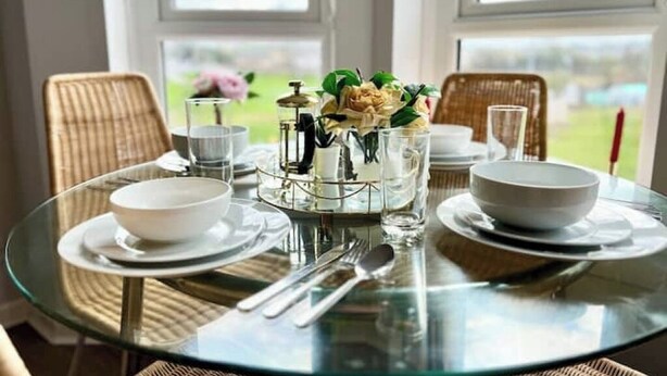 Una mesa redonda de cristal con platos, cuencos y cubiertos blancos, frente a una ventana con vistas a zonas verdes.