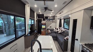 Interior compacto de um veículo recreativo, com área de refeições, sofá, televisão e escada.