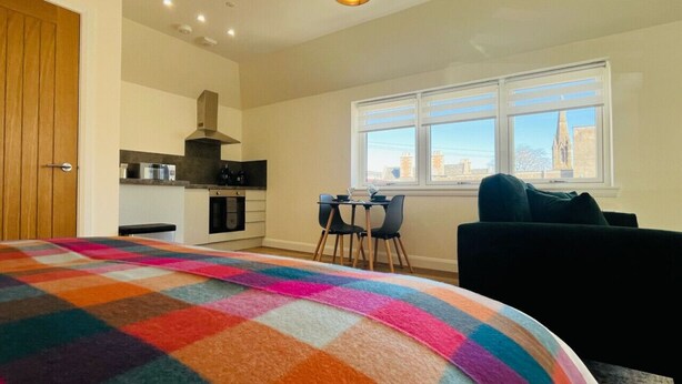 A bedroom with a colorful blanket, a dining area with chairs, a kitchen with appliances, and a window with a view. - Viewforth Studio No.3 (Fort William)