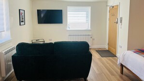 A compact room with a dark green sofa, a flat-screen TV mounted on the wall, a wooden door, and a window with blinds. - Viewforth Studio No.3 (Fort William)
