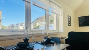 A dining area with a table set for two, a window with a view of buildings and hills, and a dark-colored sofa in the background. - Viewforth Studio No.3 (Fort William)
