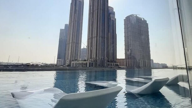 Une piscine avec des chaises modernes, entourée de grands immeubles et surplombée par un ciel dégagé.