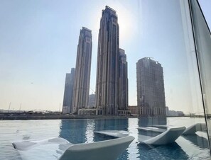 A view of a swimming pool with modern chairs, surrounded by tall buildings and a clear sky.
